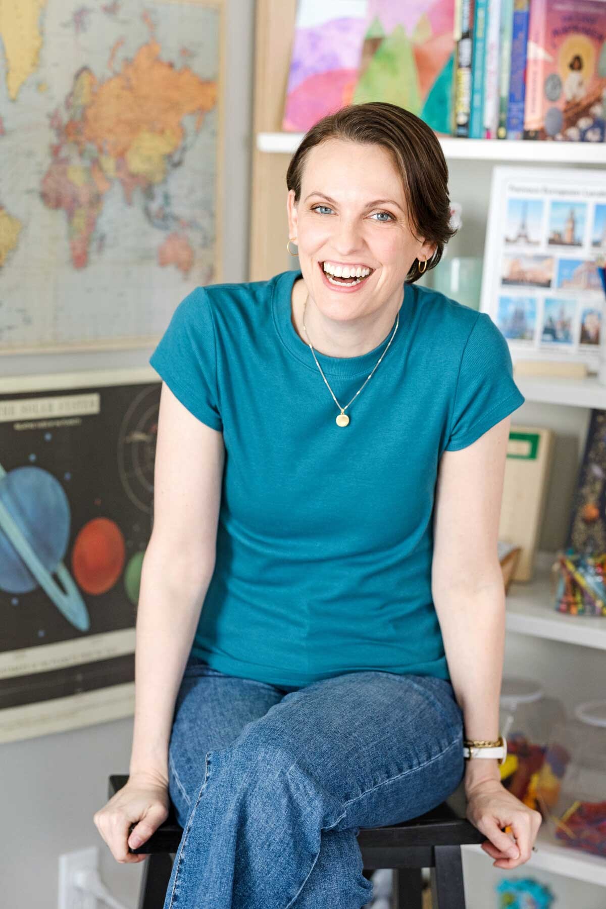Image is of a woman in a turquoise shirt sitting on a black stool with a map and a solar system poster behind her.