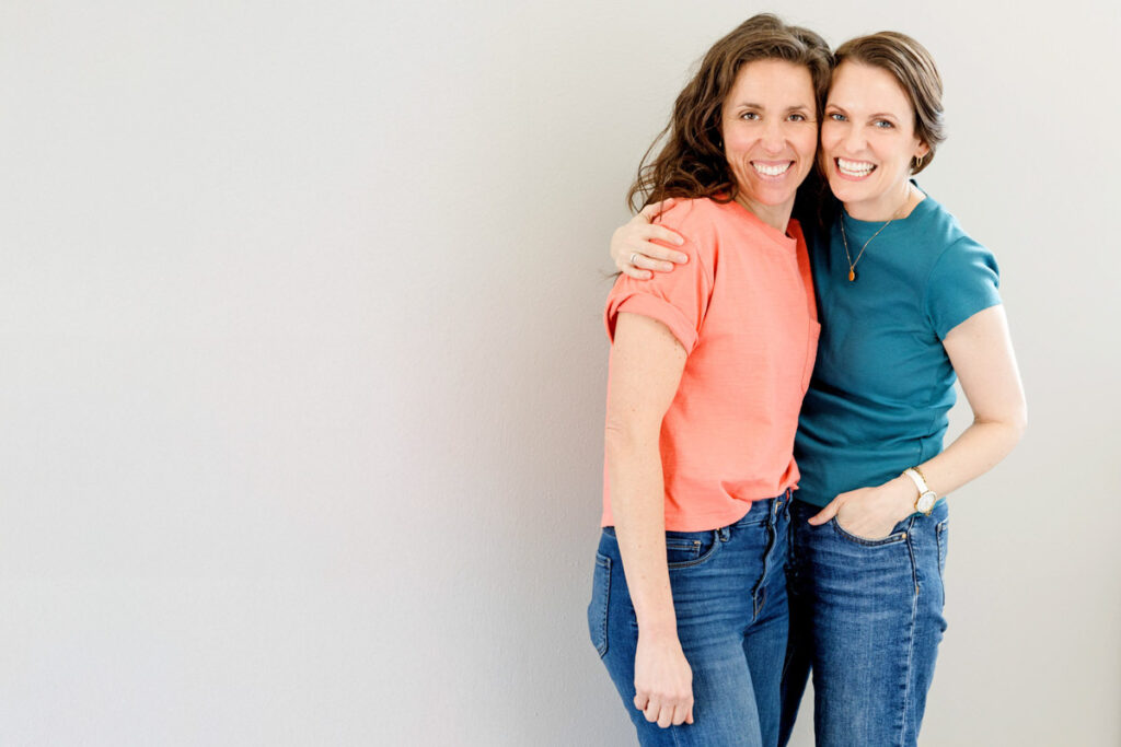 Image of two woman close together standing at a gray wall. One woman is wearing orange. The other woman is in turquoise.