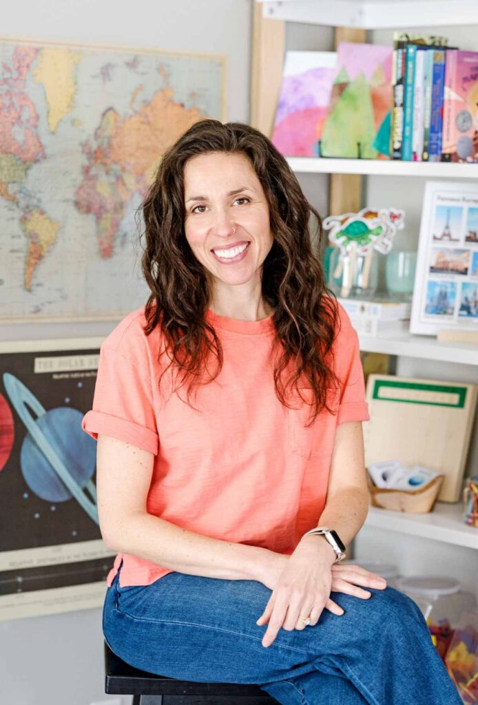 Image is of a woman in an orange shirt with long brown curly hair. She is sitting in front of a map and a solar system poster.