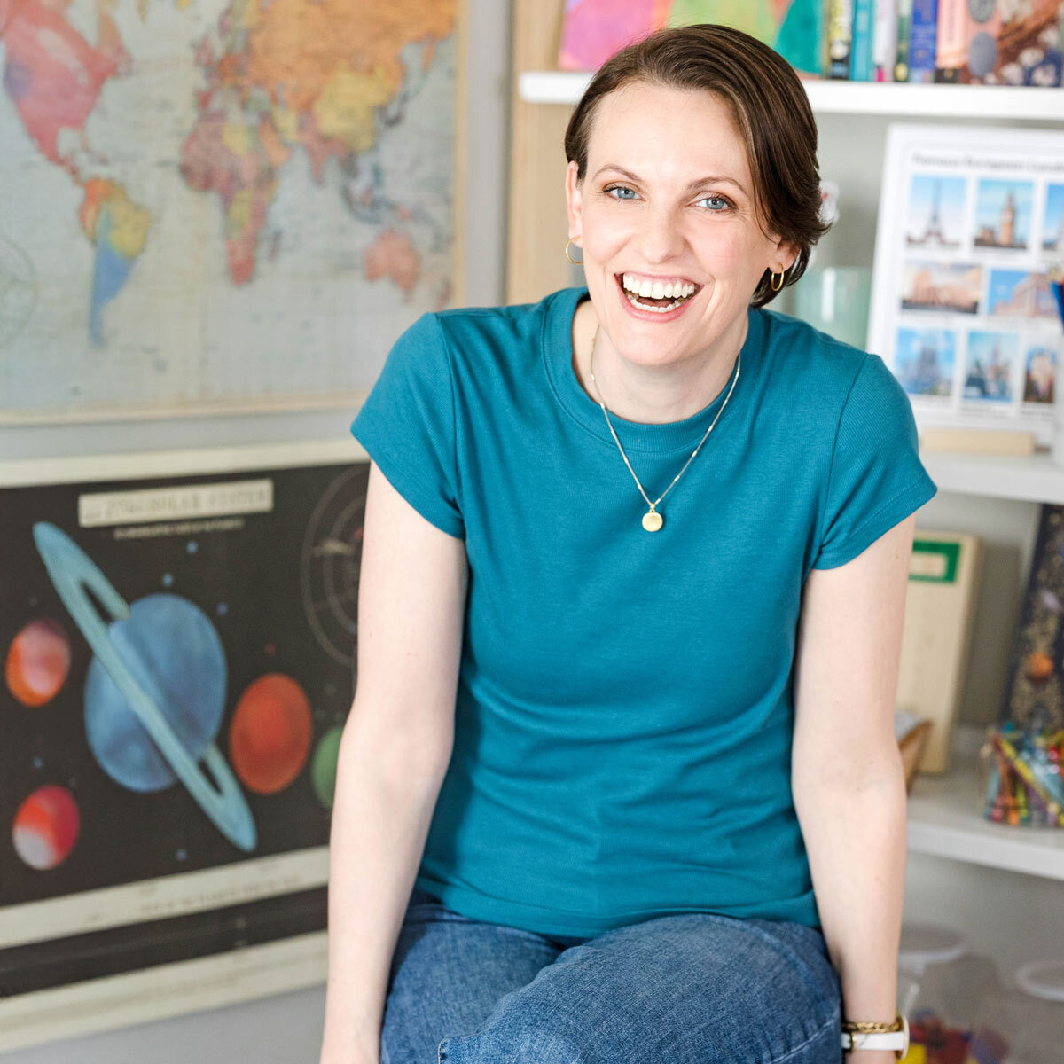 Image is of a woman in a turquoise shirt sitting on a black stool with a map and a solar system poster behind her.
